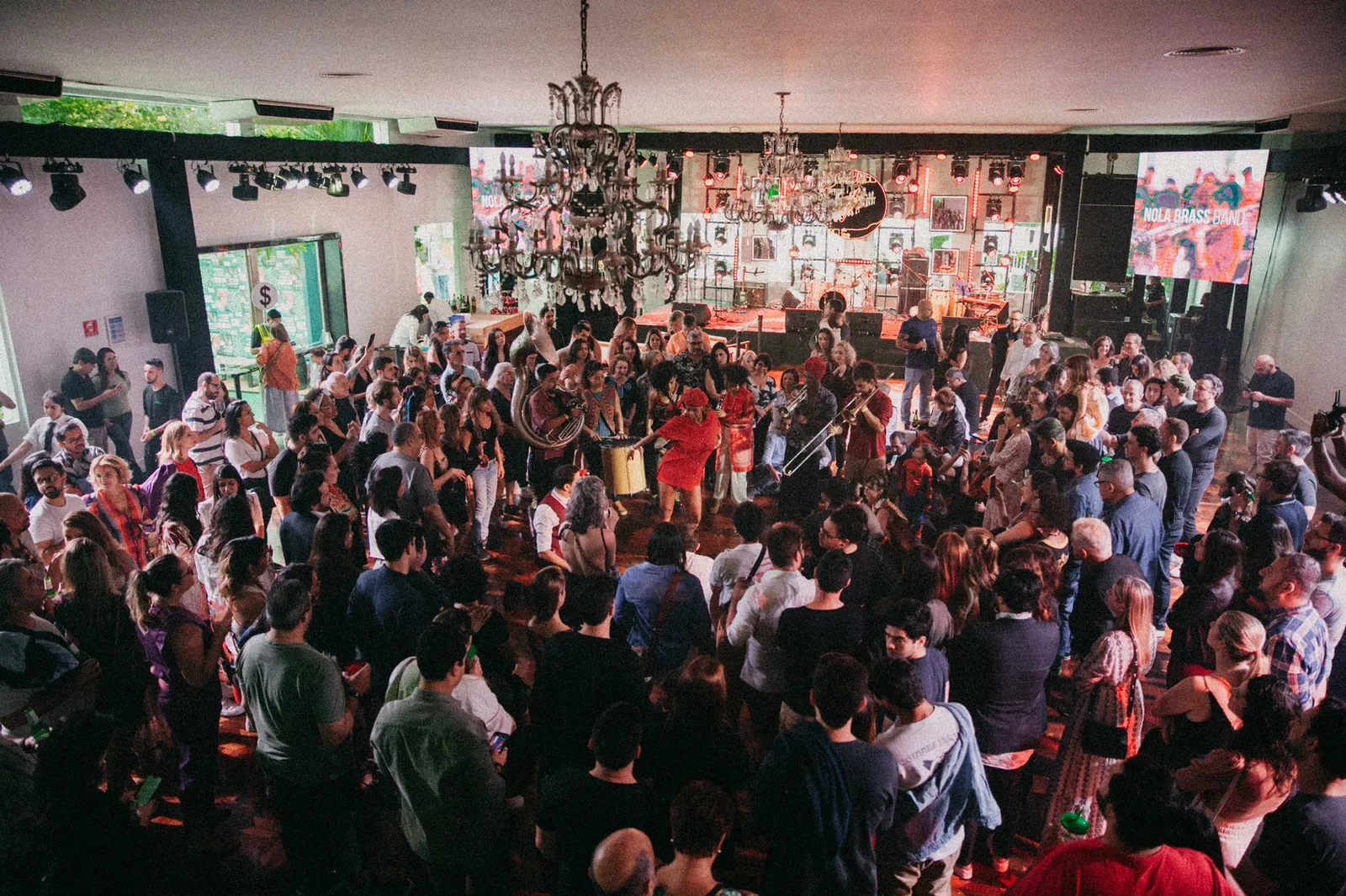 Banda de metais se apresenta cercada pelo público em um dos salões da Jazz Mansion. A imagem evidencia a proximidade entre artistas e espectadores, característica da experiência imersiva do festival.
