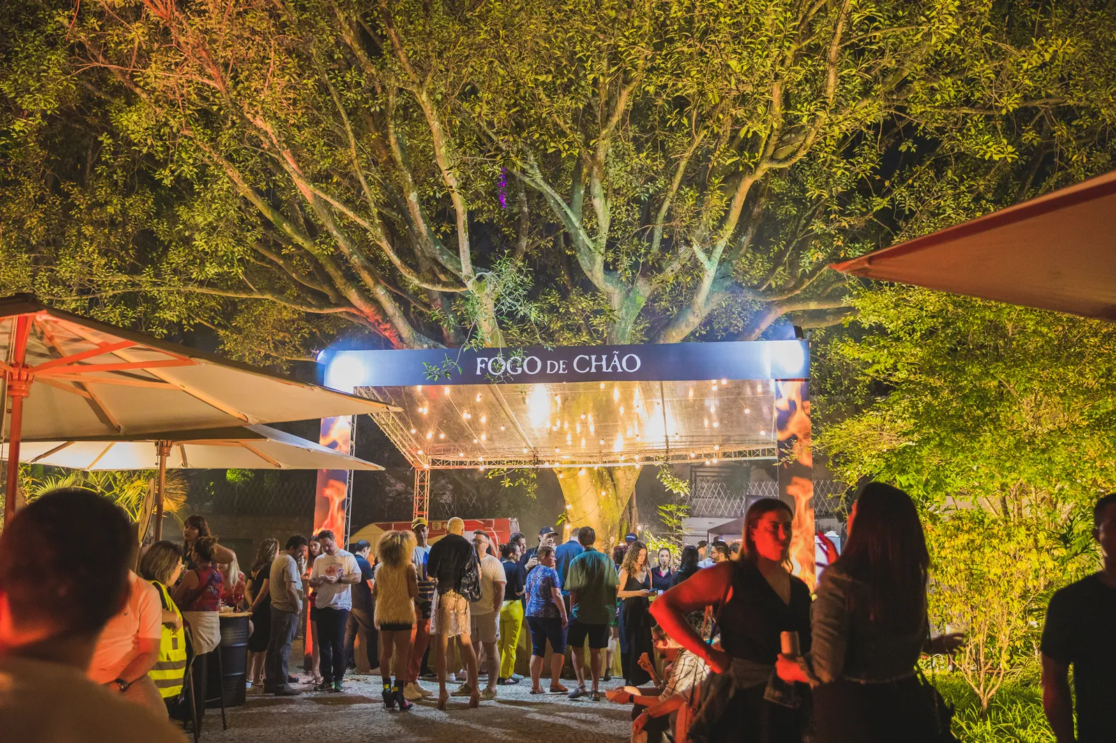 Visitantes circulam pela área gastronômica ao ar livre da Jazz Mansion, iluminada por grandes árvores e estruturas de atendimento. A imagem destaca um dos ambientes de convivência e alimentação do festival.