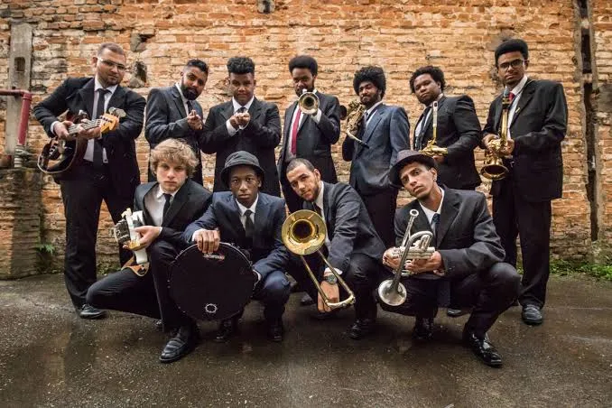 Integrantes da Nova Malandragem posam com trompetes, trombones, saxofones e outros instrumentos de sopro. A imagem apresenta a brass band responsável por levar a energia das marching bands à programação da Jazz Mansion.