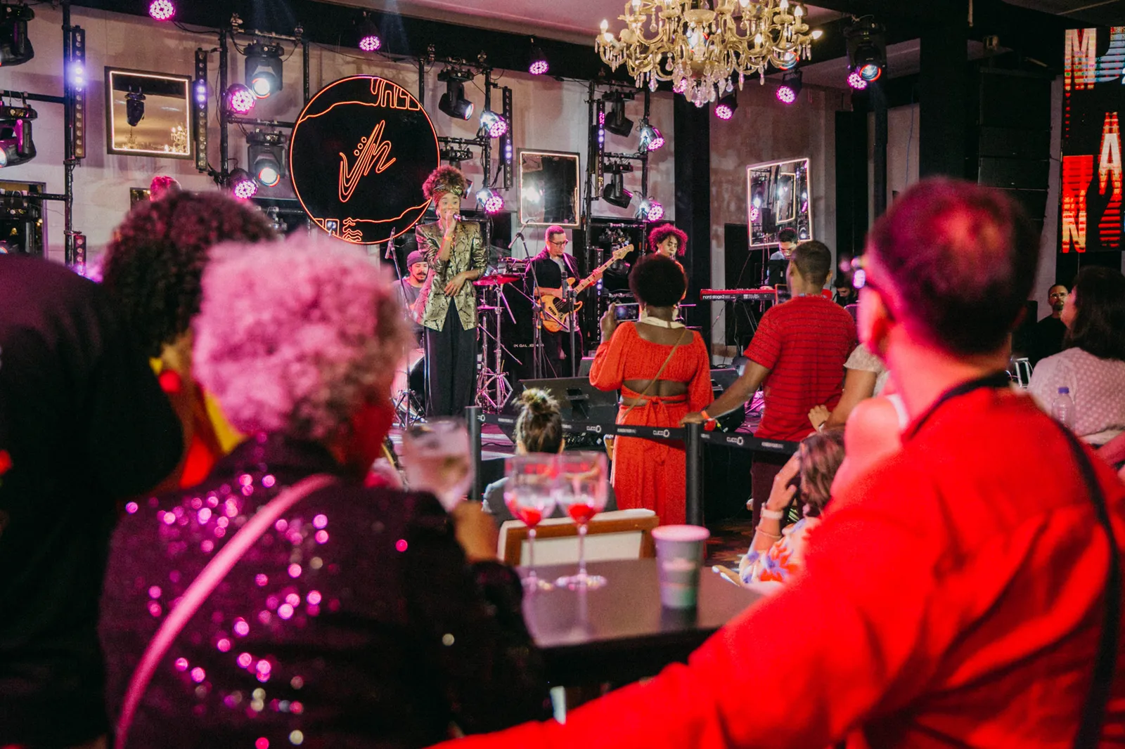 Artistas se apresentam em um palco interno enquanto o público acompanha o show sentado e em pé. A imagem destaca um dos espaços dedicados à programação musical da Jazz Mansion.