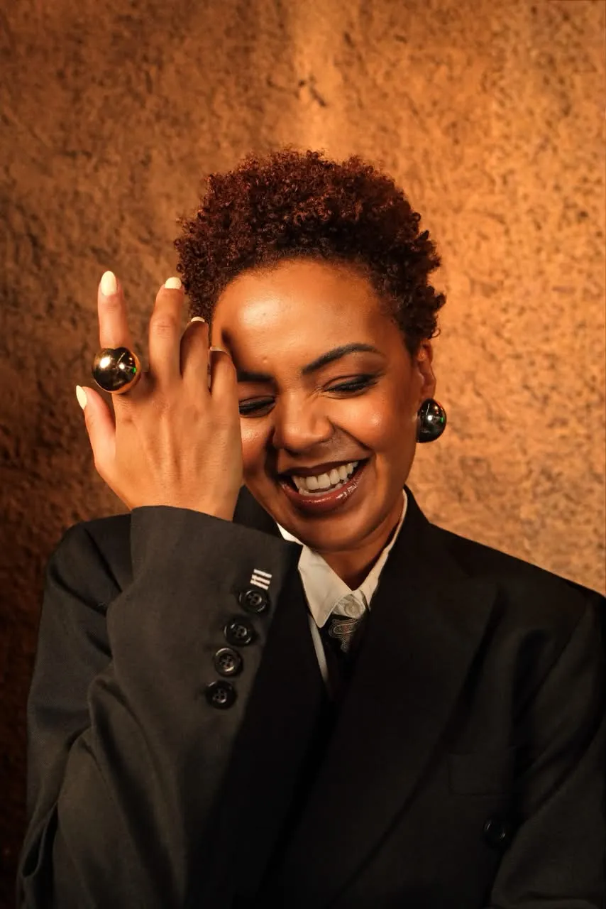 Artista sorri para a câmera usando blazer preto, camisa branca e acessórios metálicos. A imagem apresenta uma das atrações da programação da Jazz Mansion e reforça a diversidade de artistas do festival.