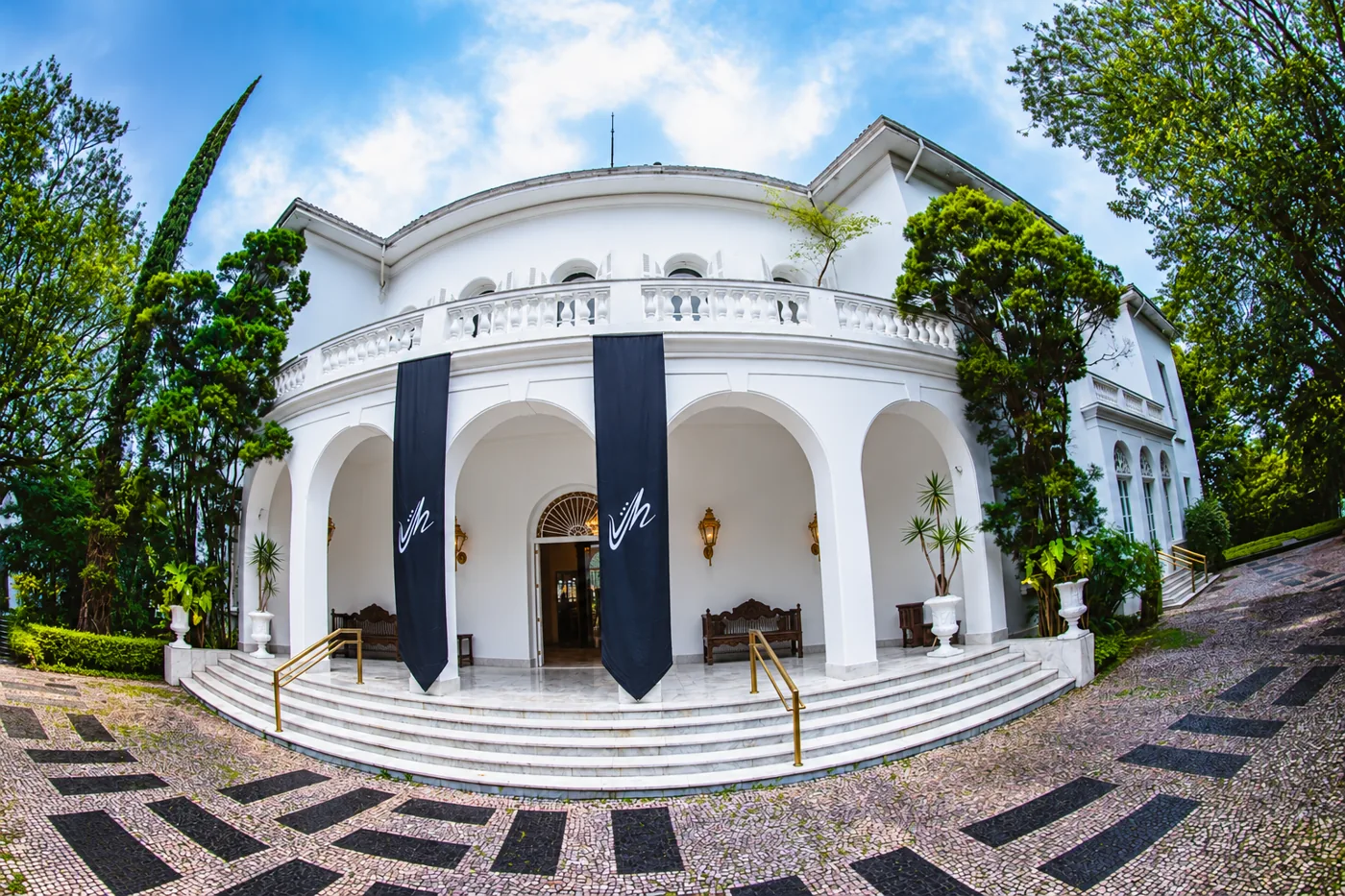 Fachada branca da mansão do Festival Jazz Mansion, com as flâmulas pretas do brasão na entrada